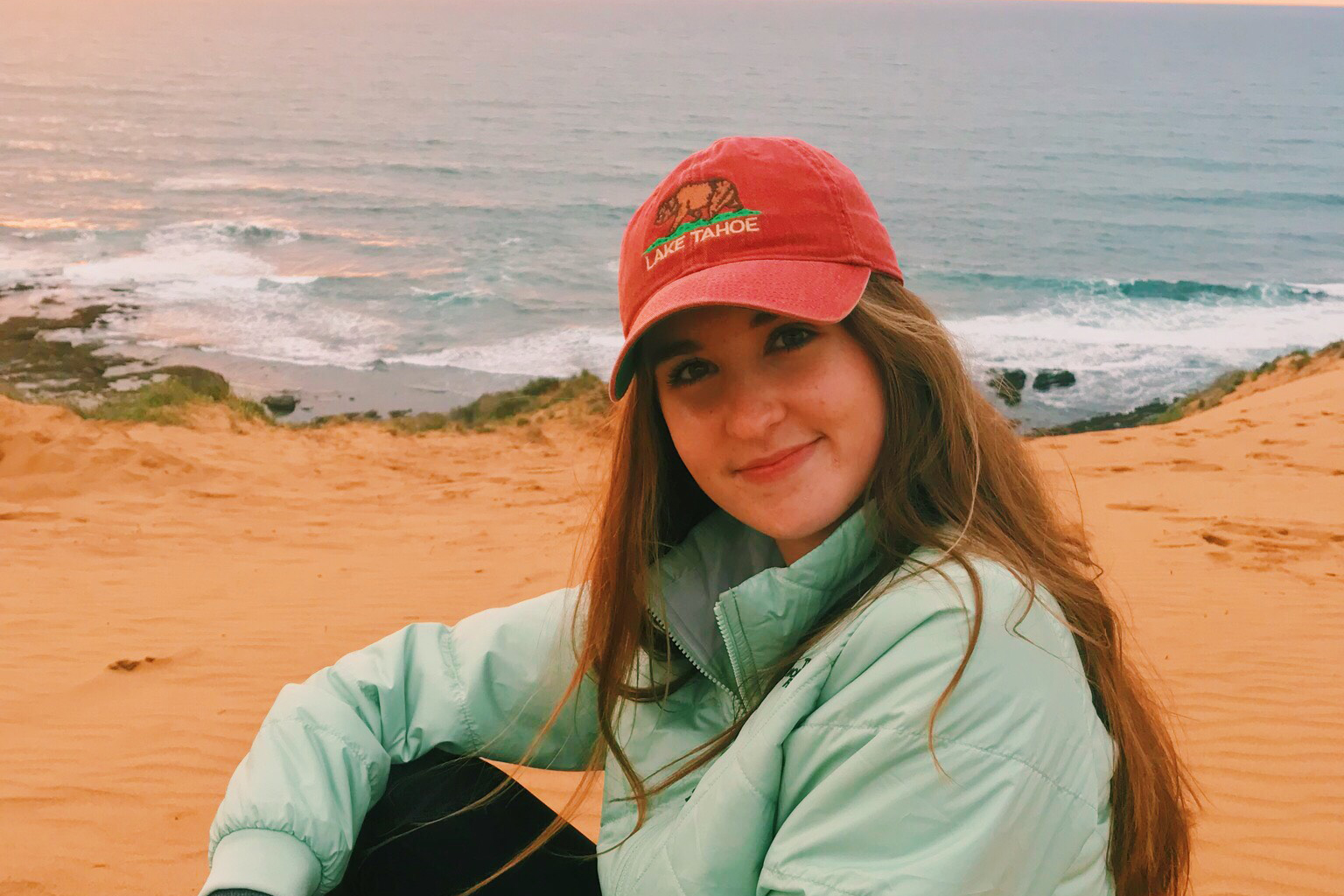 Erin Coltharp smiles in front of an ocean sunset wearing a blue jacket and red Lake Tahoe hat
