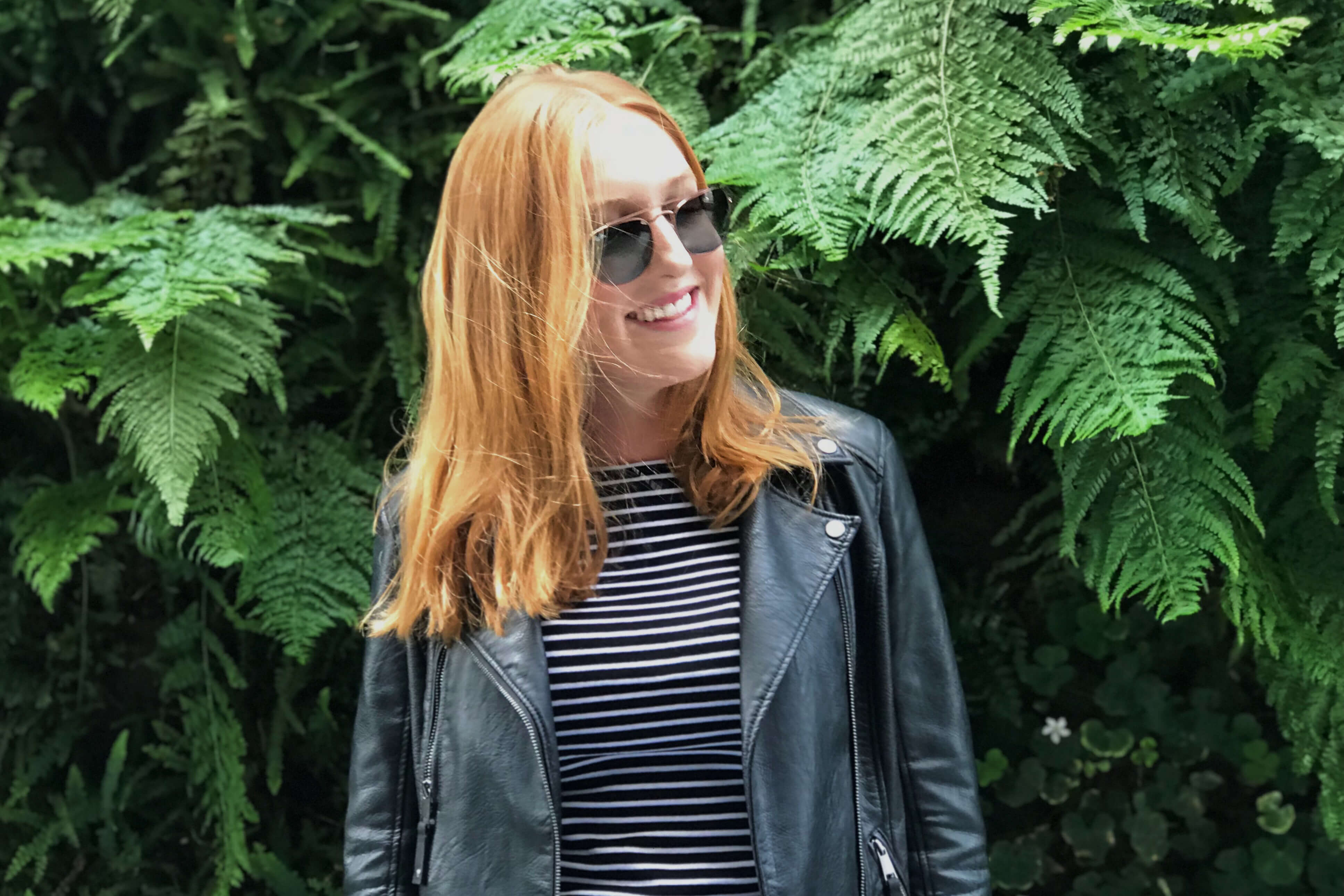 Sarah Downing smiles in front of ferns while wearing sunglasses, a leather jacket, and a striped shirt