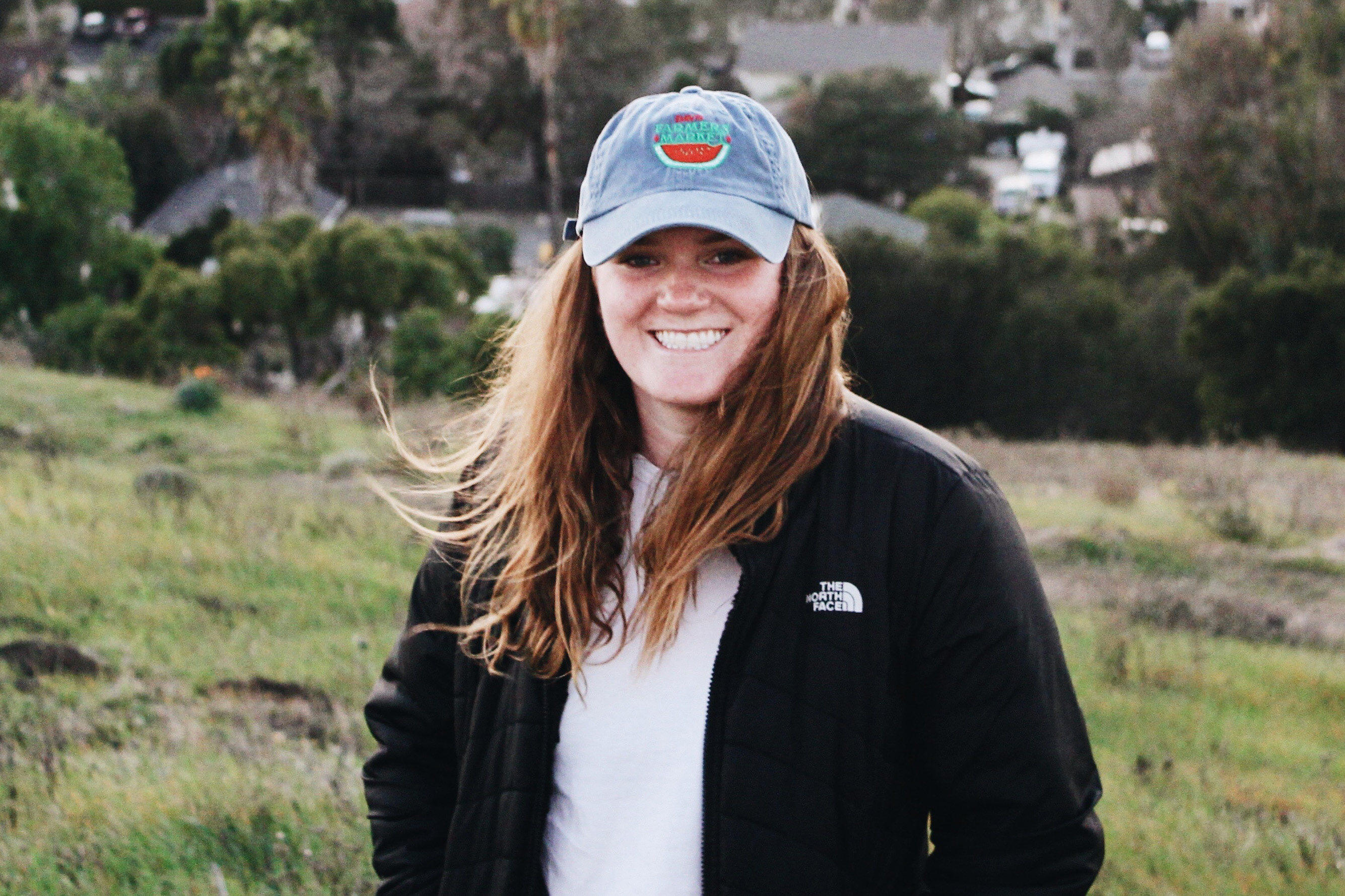 Molly Eichten smiles wearing a blue hat with a watermelon and a black North Face jacket in front of green brush and trees 